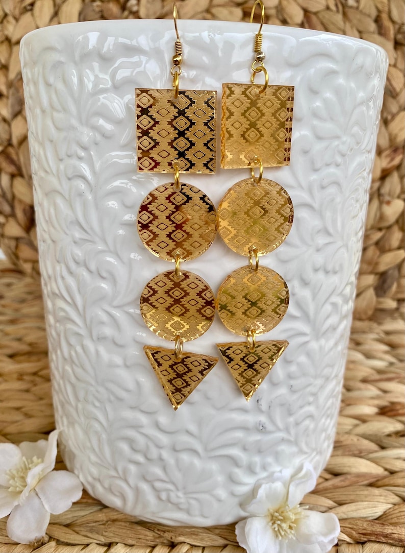 May include: A pair of gold-toned dangle earrings featuring a vertical arrangement of geometric shapes. Each earring consists of a square, two circles, and a triangle, all etched with a repeating diamond pattern and suspended from gold-toned hooks.