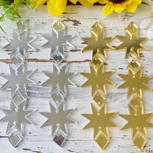 May include: A collection of star-shaped acrylic charms arranged in rows. The charms feature a reflective mirror finish in silver and gold tones. Each star has a small diamond-shaped attachment at the top and bottom, with pre-drilled holes for hanging.