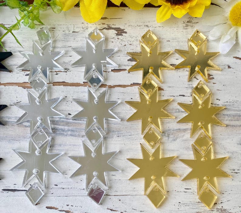 May include: A collection of star-shaped acrylic charms arranged in rows. The charms feature a reflective mirror finish in silver and gold tones. Each star has a small diamond-shaped attachment at the top and bottom, with pre-drilled holes for hanging.