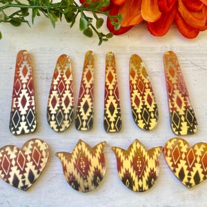 May include: A collection of ten acrylic jewelry pendants featuring a warm-toned geometric pattern in shades of orange, red, and black. The set includes six elongated teardrop shapes and four smaller pieces shaped like hearts and stylized tulips.