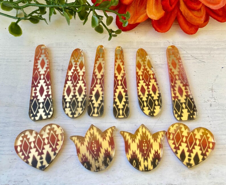 May include: A collection of ten acrylic jewelry pendants featuring a warm-toned geometric pattern in shades of orange, red, and black. The set includes six elongated teardrop shapes and four smaller pieces shaped like hearts and stylized tulips.