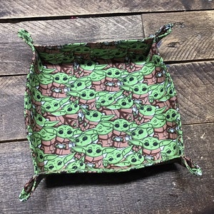 May include: A green and brown fabric tray with a repeating pattern of a cartoon character holding a cup. The tray has four corners that fold up to create a small, square-shaped container.