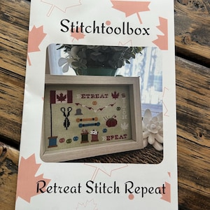 Puede incluir: Un papel blanco con el texto "Stitchtoolbox" y "Retreat Stitch Repeat" en negro. El papel tiene una ventana que revela un bordado enmarcado con una bandera canadiense y las palabras "Retreat Stitch Repeat". El fondo es una mesa de madera.