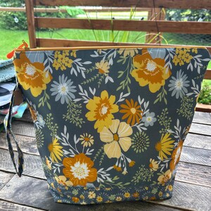 May include: A zipped pouch featuring a dark grey background with a repeating floral pattern in shades of mustard yellow, golden orange, and pale grey. The bag includes a matching fabric wrist strap and a bright orange zip pull tab at the top.