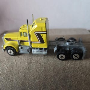 May include: A yellow die-cast semi-truck cab featuring a black and white stripe graphic on the side, silver exhaust stacks, a gray chassis, and black wheels with white rims. The vehicle is shown from a side profile view, highlighting the cab design.