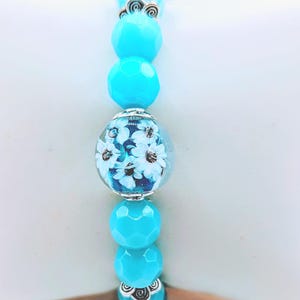 May include: A close-up of a bracelet featuring turquoise faceted beads and a clear bead with white floral design. The bracelet has silver-colored accents. The beads are strung together, creating a decorative accessory.