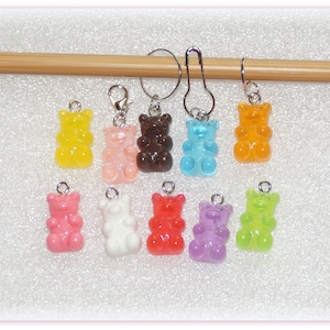 Könnte beinhalten: Ein Set mit 10 bunten Gummibärchen-Maschenmarkierern, jeder mit einer silbernen Schlaufe und einem silbernen Ring. Die Marker sind in einer Reihe auf einer Holzoberfläche angeordnet.