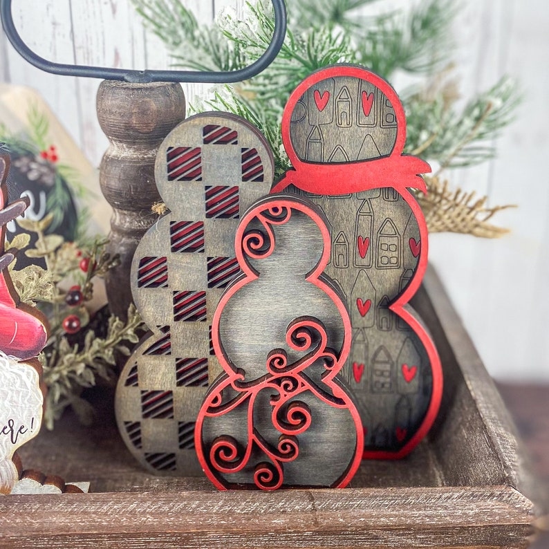 May include: Three wooden snowman ornaments with red and black paint. The snowman on the left has a checkered pattern, the middle snowman has a swirl design, and the snowman on the right has a pattern of hearts and houses.