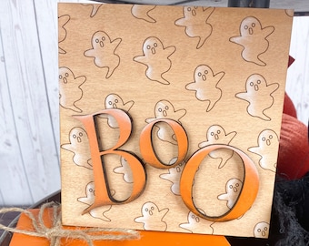 Może przedstawiać: Drewniany znak z wzorem duchów i napisem "Boo" w pomarańczowych literach. Znak idealnie nadaje się do dekoracji na Halloween.