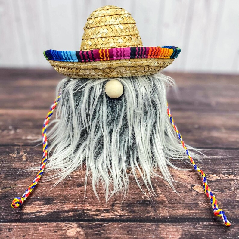 May include: A white and gray fuzzy gnome wearing a straw sombrero with a colorful band. The gnome has a wooden nose and a long, braided cord.