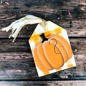 以下が含まれることがあります： 黄色と白のギンガムチェック柄の、木製の南瓜の形をしたタグ。タグには、吊り下げ用の麻紐のループが付いており、黒の茎が付いたオレンジ色の南瓜が描かれています。