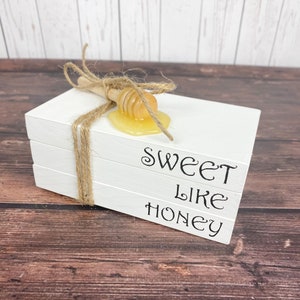 May include: Three white stacked books with the text "Sweet Like Honey" on the front. A wooden honey dipper with honey dripping from it is on top of the books.