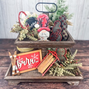May include: A two-tiered wooden tray with a rustic finish, decorated with Christmas-themed wooden ornaments. The top tier features a gnome with a red hat, a snowman, and a sign that says "Oh dear, world is here!" The bottom tier features a sign that says "burr..." a wooden sled, and a red heart ornament. The tray is surrounded by greenery and red berries.