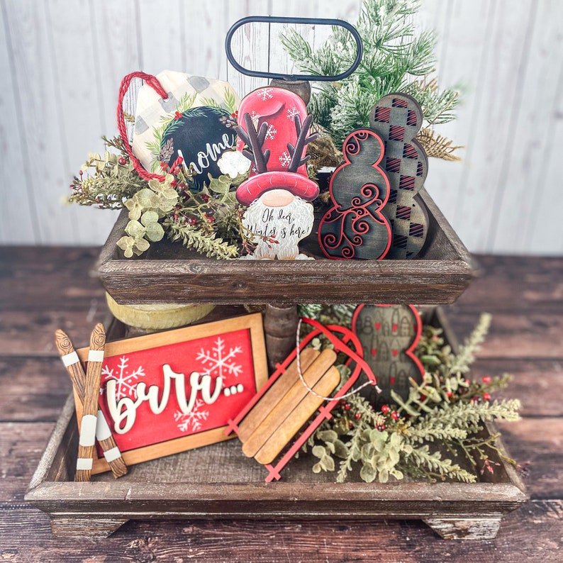 May include: A two-tiered wooden tray with a rustic finish, decorated with Christmas-themed wooden ornaments. The top tier features a gnome with a red hat, a snowman, and a sign that says "Oh dear, world is here!" The bottom tier features a sign that says "burr..." a wooden sled, and a red heart ornament. The tray is surrounded by greenery and red berries.