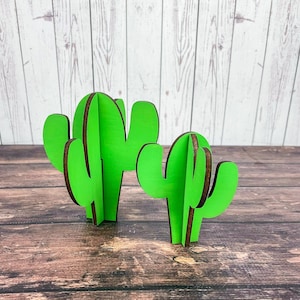 May include: Two green wooden cactus cutouts. The larger cactus is on the left and the smaller cactus is on the right. Both cacti have a brown wood grain.