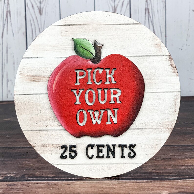 Może przedstawiać: Białe drewniane koło z namalowanym na nim czerwonym jabłkiem. Jabłko ma napisane na sobie słowa "PICK YOUR OWN". Pod jabłkiem znajduje się tekst "25 CENTS".