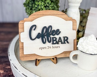 May include: A wooden sign with the words "Coffee Bar" in black lettering and the text "open 24 hours" below. The sign is on a white stand and is sitting on a white surface.