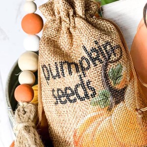 以下が含まれることがあります： ひもで閉じた小さな麻袋。袋には、かぼちゃの絵と「pumpkin seeds」の文字が描かれています。