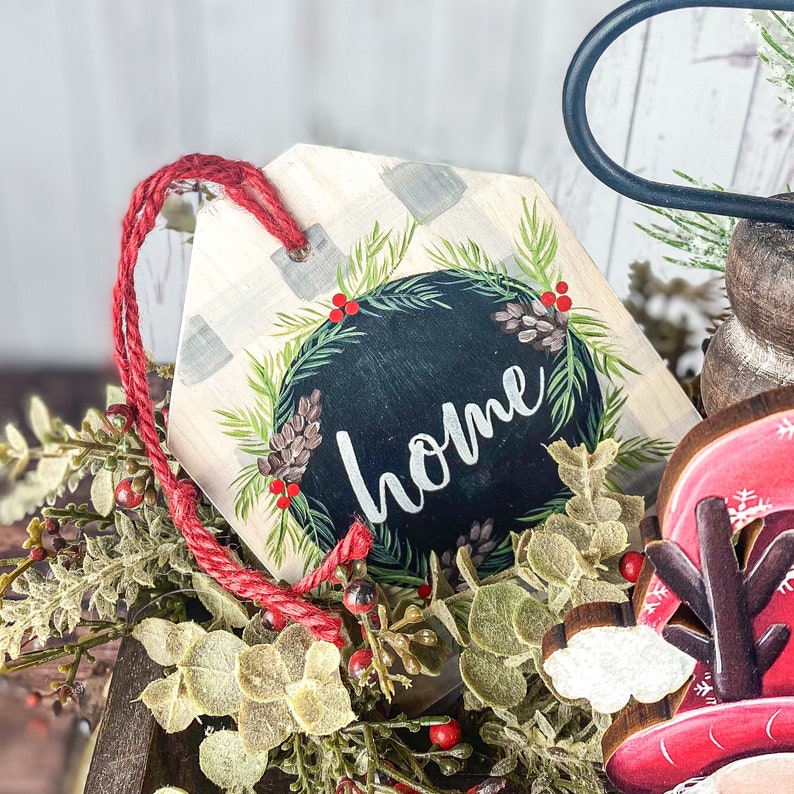 May include: A wooden sign with a white background and a black chalkboard circle with the word "home" written in white. The sign is decorated with a green wreath with red berries and pine cones.