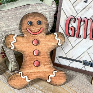May include: A wooden gingerbread man ornament with white icing details and red button accents. The gingerbread man is smiling and has brown eyes.