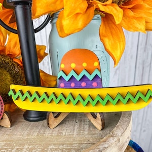 May include: A colorful wooden sombrero with a green, yellow, orange, and purple design. The sombrero is on a wooden stand.