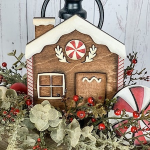 May include: A wooden gingerbread house decoration with white trim, a red and white candy accent, and a chimney. The house is surrounded by greenery and red berries, creating a festive holiday scene.