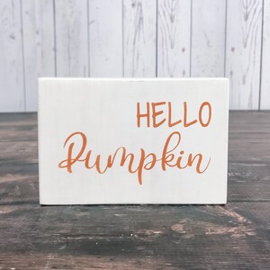 以下が含まれることがあります： オレンジ色の文字で「HELLO PUMPKIN」と書かれた白い木製の看板です。