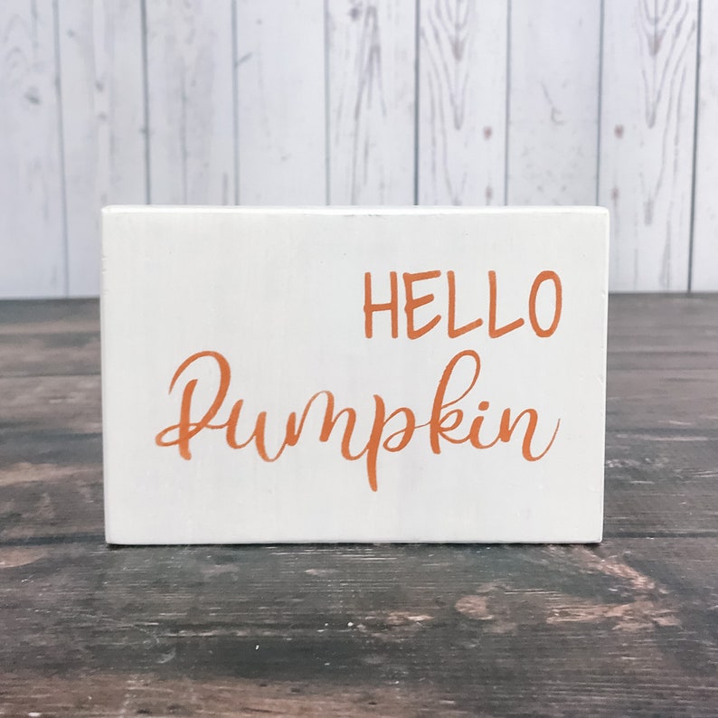 以下が含まれることがあります： オレンジ色の文字で「HELLO PUMPKIN」と書かれた白い木製の看板です。