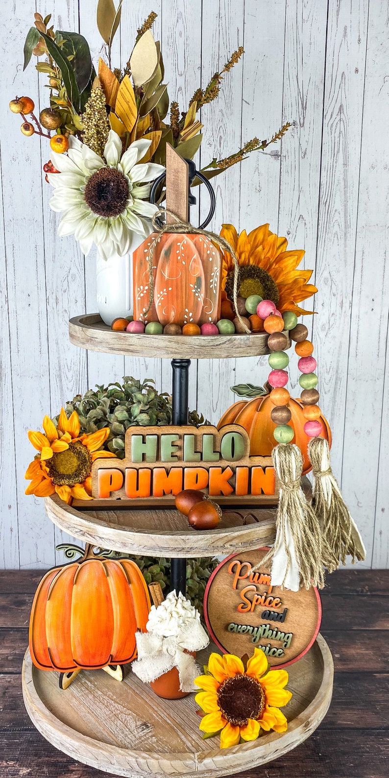 May include: A three-tiered wooden tray with fall decorations. The top tier has a white vase with a sunflower and greenery, a wooden pumpkin with orange and brown paint, and a beaded garland in pink, brown, and green. The middle tier has a wooden sign that reads 'HELLO PUMPKIN' in green and orange letters, a sunflower, and a brown tassel. The bottom tier has a wooden pumpkin with orange paint, a white jar with a white lid, a wooden sign that reads 'Pumpkin Spice and Everything Nice' in orange and brown letters, and a sunflower.