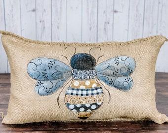 May include: A beige burlap pillow with a hand-painted blue and yellow bee design. The bee has black and white polka dots and a checkered pattern on its body.