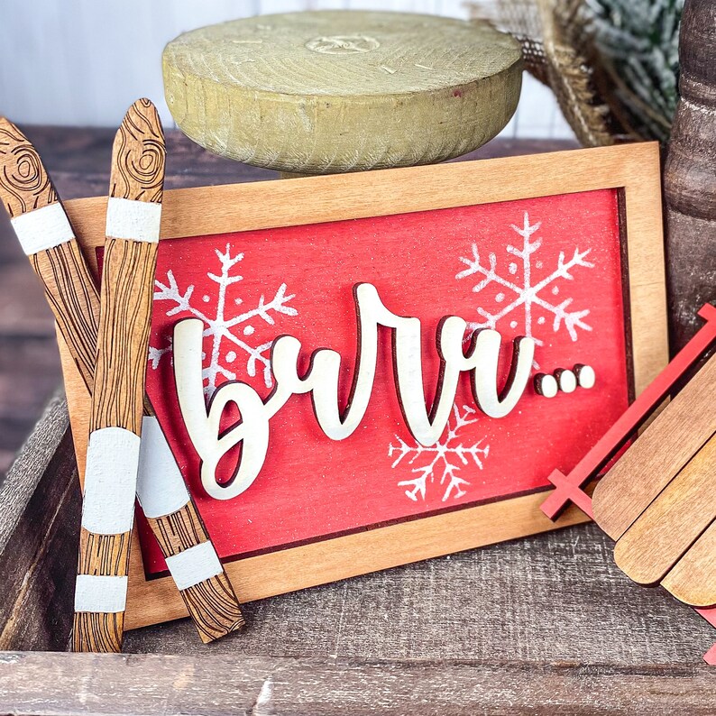 May include: Red wooden sign with the word "burr..." in white painted letters. The sign is decorated with white snowflakes. Two wooden skis with white stripes are leaning against the sign.