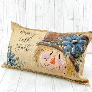 May include: Rectangular burlap pillow with a painted scarecrow design. The scarecrow wears a brown hat adorned with blue flowers and green leaves. The pillow has the text "Happy Fall Y'all" in black script.