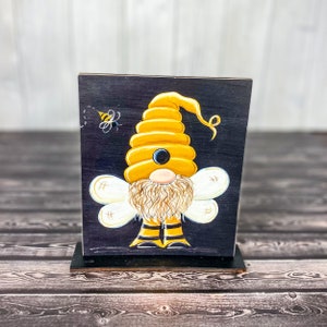 May include: A painted wooden gnome with a yellow honeycomb hat and white wings. The gnome is standing on a black background with a small bee flying nearby.