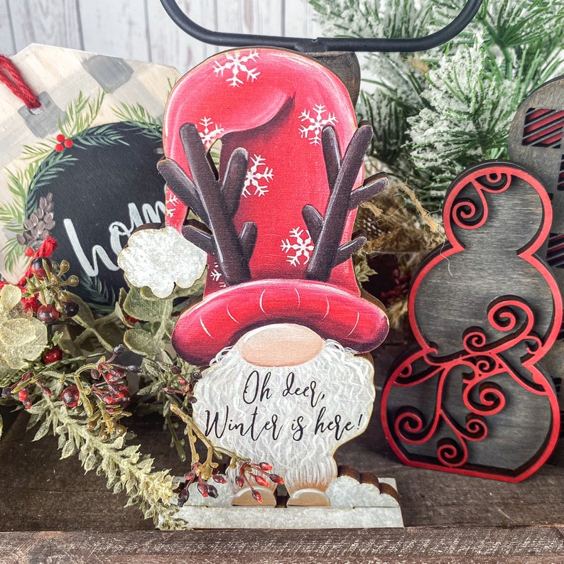 May include: A wooden gnome figurine with red and white hat, antlers, and a white beard. The gnome is standing on a white base and has the text "Oh deer, Winter is here!" on its belly.