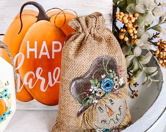 Puede incluir: Una pequeña bolsa de arpillera con una cara de espantapájaros pintada y una corona de flores azul y blanca. La bolsa está sobre una superficie blanca con una calabaza pintada en el fondo. La calabaza tiene las palabras "Happy Harvest" pintadas en ella.