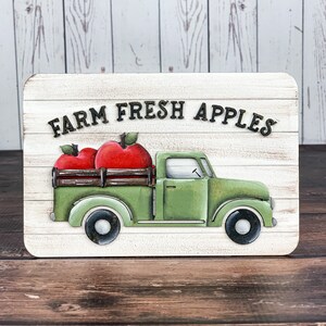 Może przedstawiać: Biały drewniany znak z napisem "FARM FRESH APPLES" czarnymi literami. Na znaku namalowany jest zielony, zabytkowy samoch&oacute;d dostawczy z drewnianą skrzynią ładunkową. Samoch&oacute;d przewozi dwa czerwone jabłka.
