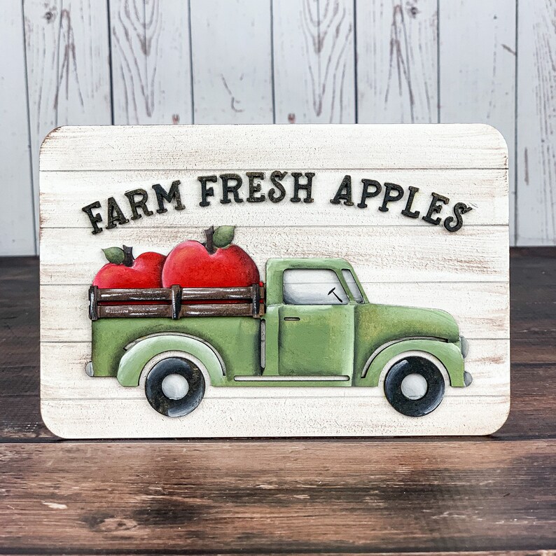 Może przedstawiać: Biały drewniany znak z napisem "FARM FRESH APPLES" czarnymi literami. Na znaku namalowany jest zielony, zabytkowy samoch&oacute;d dostawczy z drewnianą skrzynią ładunkową. Samoch&oacute;d przewozi dwa czerwone jabłka.