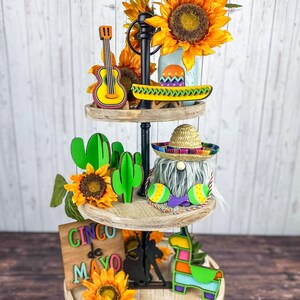 May include: A three-tiered wooden tiered tray decorated with a Cinco de Mayo theme. The top tier features a yellow guitar, a green and yellow canoe, and sunflowers. The middle tier has a gnome wearing a sombrero, green cacti, and sunflowers. The bottom tier has a sign that says "Cinco de Mayo", a black silhouette of a person riding a horse, a llama, and sunflowers.