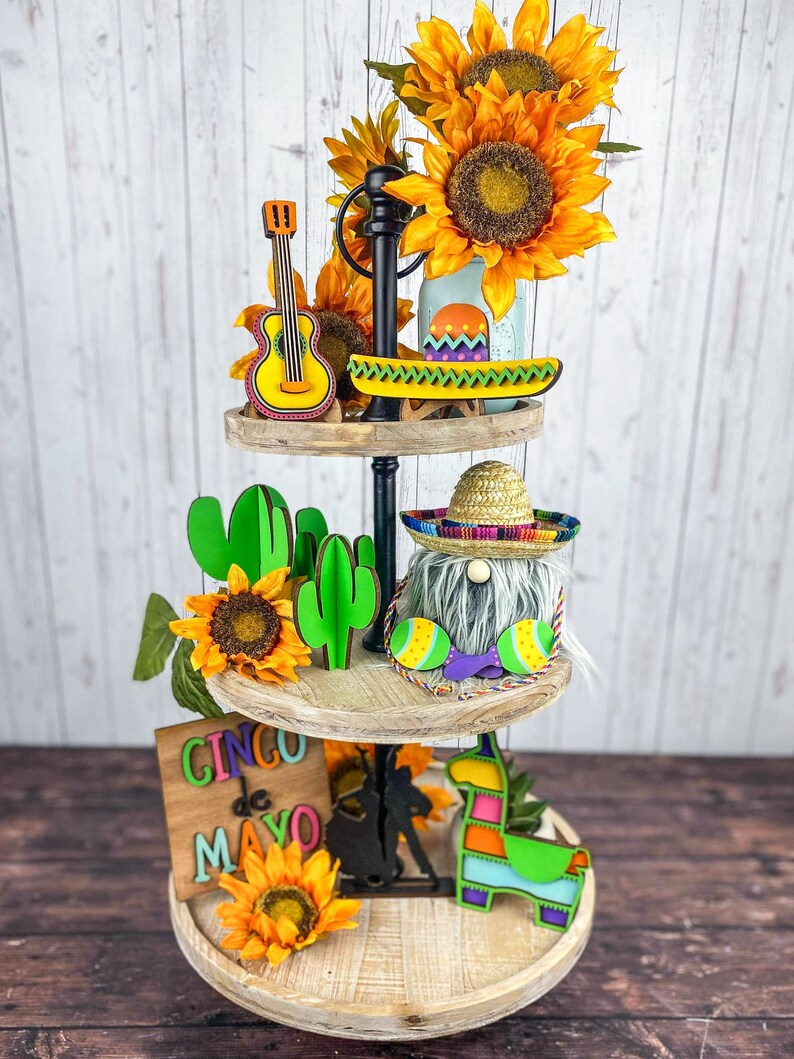 May include: A three-tiered wooden tiered tray decorated with a Cinco de Mayo theme. The top tier features a yellow guitar, a green and yellow canoe, and sunflowers. The middle tier has a gnome wearing a sombrero, green cacti, and sunflowers. The bottom tier has a sign that says "Cinco de Mayo", a black silhouette of a person riding a horse, a llama, and sunflowers.