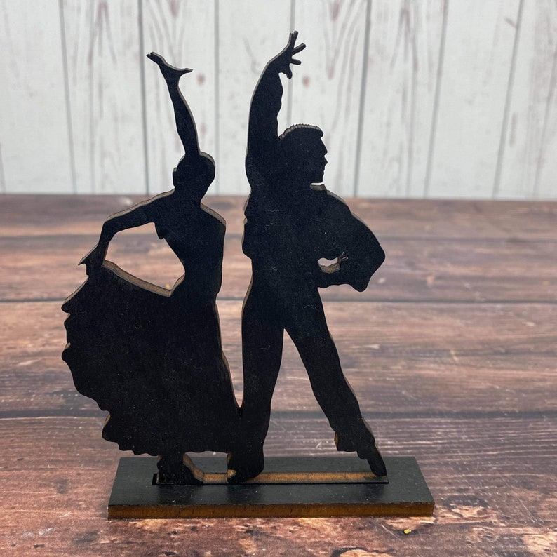 May include: Black silhouette of a couple dancing, the woman in a flowing skirt and the man in a suit, on a wooden base.