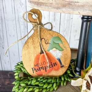 May include: A wooden cutting board with a painted gnome and pumpkin. The pumpkin is orange and has the words "Hello Pumpkin" painted on it. The gnome is wearing a green hat and has a white beard.
