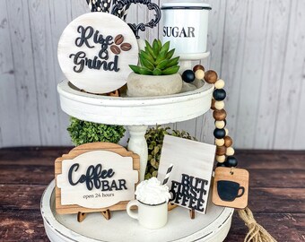May include: A white tiered tray with three levels, decorated with coffee-themed signs, a small white sugar container, a succulent plant, and a black and white striped straw. The signs read "Rise & Grind", "Coffee Bar open 24 hours", and "But First Coffee".
