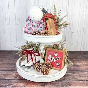 May include: A two-tiered white tiered tray decorated for winter with a red and white knit hat, a red wooden sled, pine cones, and a red sign that says "Brr".