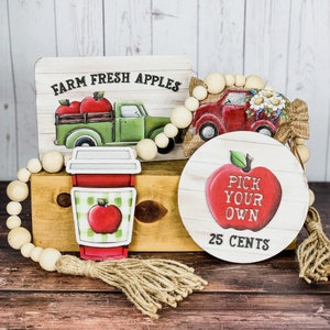 Może przedstawiać: Drewniany znak z napisem "Farm Fresh Apples" i czerwonym ciężar&oacute;wką z jabłkami z tyłu. Czerwony ciężar&oacute;wka z białymi kwiatami na g&oacute;rze. Drewniany znak z czerwonym jabłkiem i napisem "Pick Your Own 25 Cents". Czerwono-biały kubek do kawy w kratkę z czerwonym jabłkiem na nim. Drewniany naszyjnik z koralik&oacute;w.