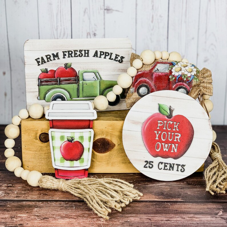 Może przedstawiać: Drewniany znak z napisem "Farm Fresh Apples" i czerwonym ciężar&oacute;wką z jabłkami z tyłu. Czerwony ciężar&oacute;wka z białymi kwiatami na g&oacute;rze. Drewniany znak z czerwonym jabłkiem i napisem "Pick Your Own 25 Cents". Czerwono-biały kubek do kawy w kratkę z czerwonym jabłkiem na nim. Drewniany naszyjnik z koralik&oacute;w.
