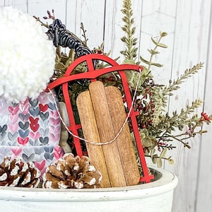 May include: A red wooden sled with a white rope, surrounded by greenery and a white pom-pom.