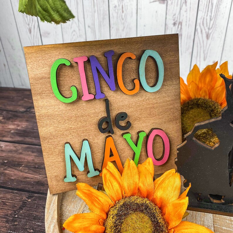May include: A wooden sign with the words "Cinco de Mayo" in colorful letters. The sign is sitting on a table with a sunflower in the foreground.