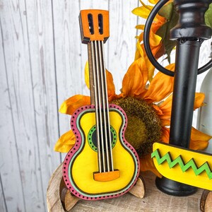 May include: A wooden cutout of a yellow guitar with pink, green, and black accents. The guitar is on a wooden stand and has a sunflower in the background.