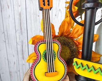 May include: A wooden decorative guitar with a yellow body, pink and green accents, and brown neck. The guitar is part of a festive display, with a sunflower and other decorative elements in the background.