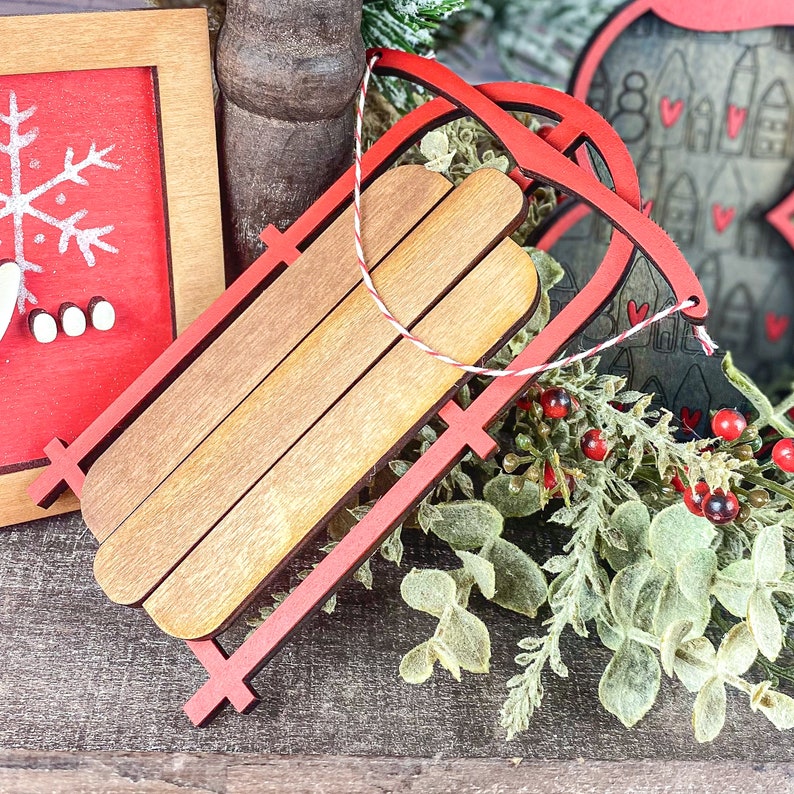 May include: A red wooden sled ornament with a brown wooden base. The ornament is decorated with a red and white string.